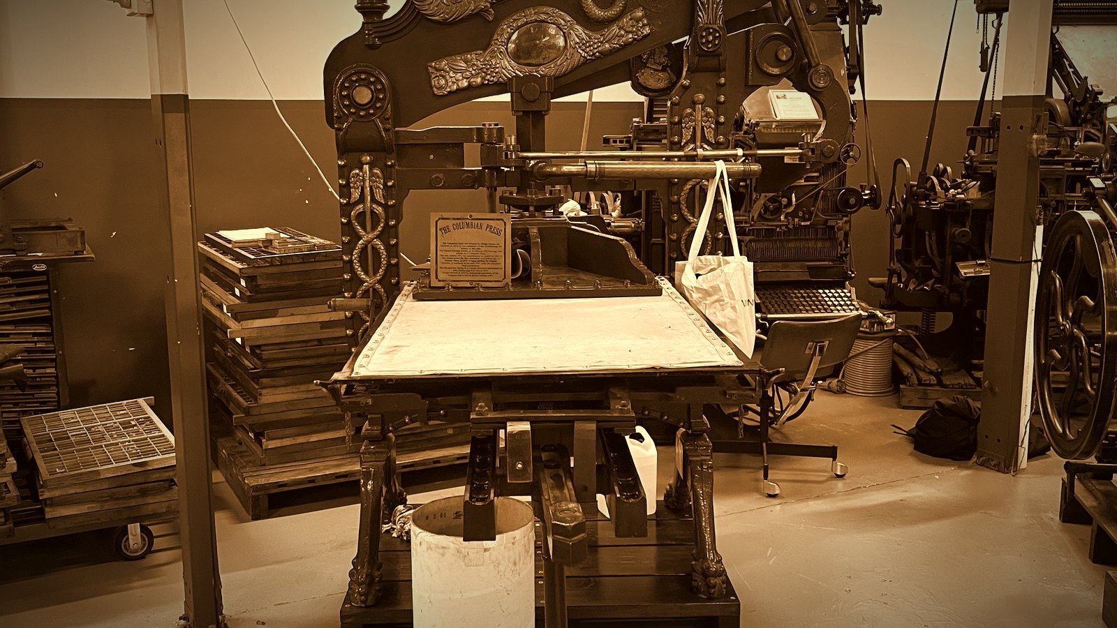 An ornate cast-iron Columbian hand printing press with trays of type beside it in a printing museum; warm sepia tones