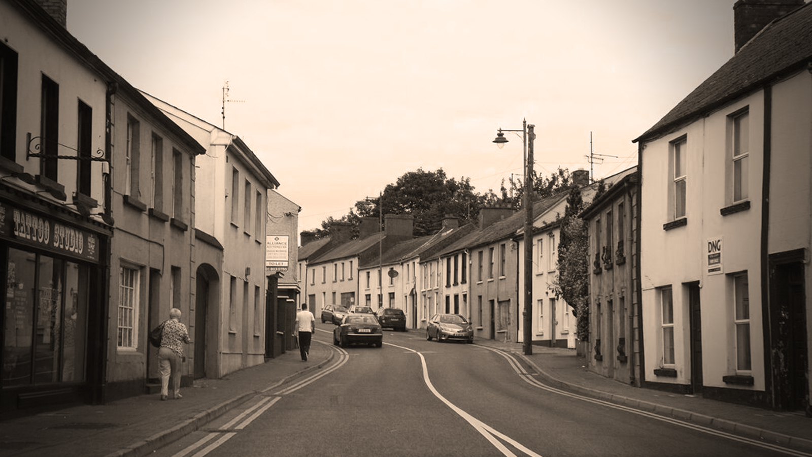 Carrick Street in Kells, a row of terraced houses and shops along a curving road, with a woman walking on the footpath; sepia tones