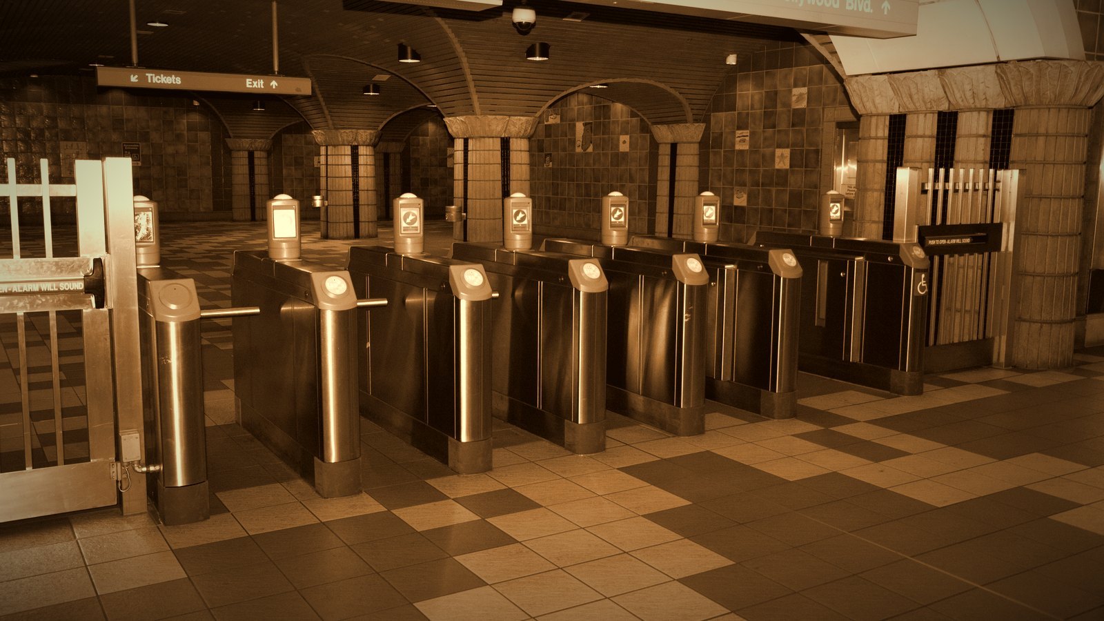 A row of metal ticket turnstiles in a tiled station hall; warm sepia tones