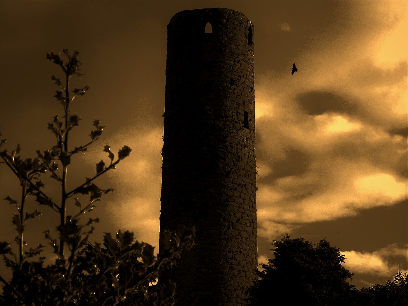 Photograph of the Round Tower in Kells, Co. Meath, seen from below against a cloudy sky, its conical cap missing; colour-toned sepia