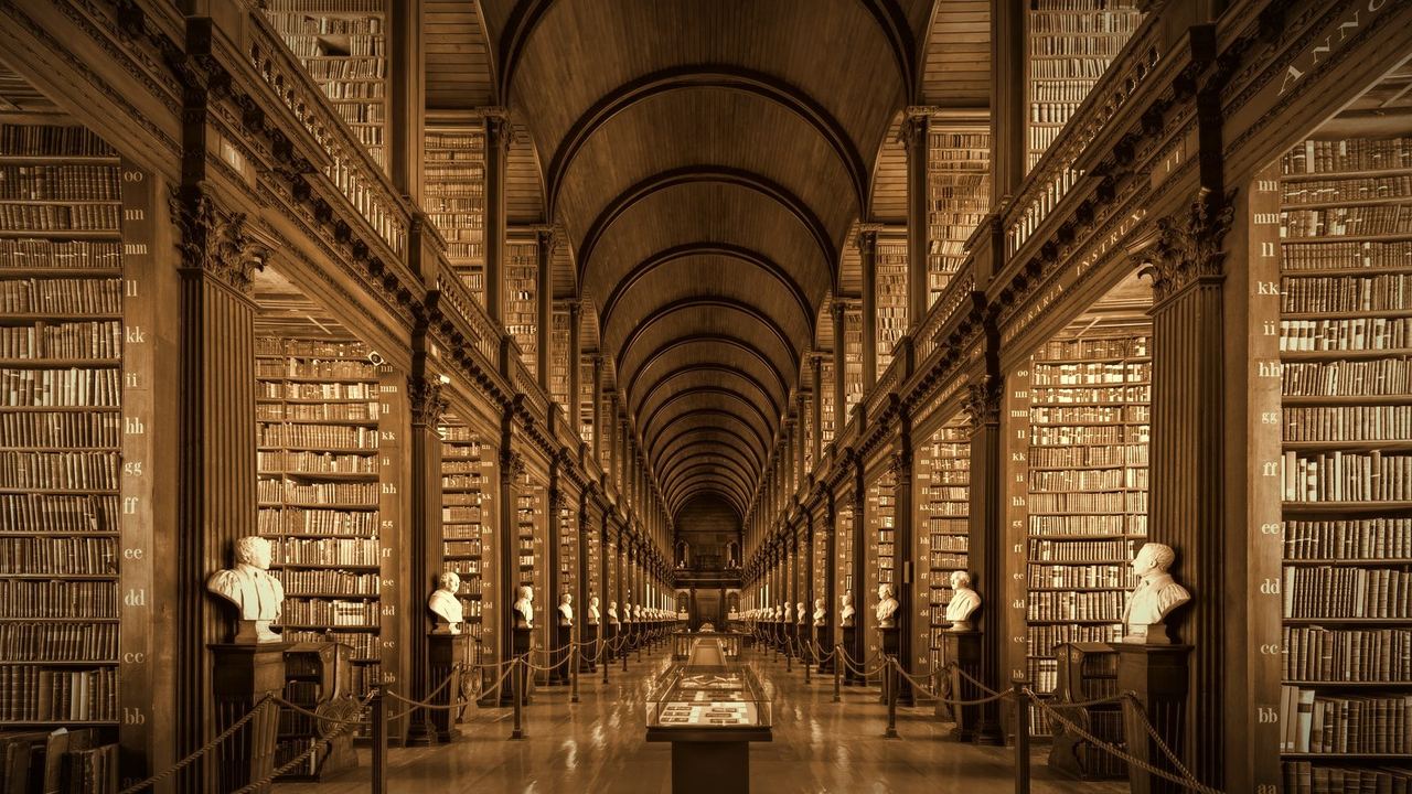 The barrel-vaulted Long Room of the Old Library at Trinity College Dublin, lined with tall bookcases and marble busts, in warm sepia tones